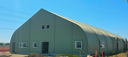 Temporary emergency housing structure with a large green, arched building, windows, and an open doorway, set on a construction site under a clear blue sky.